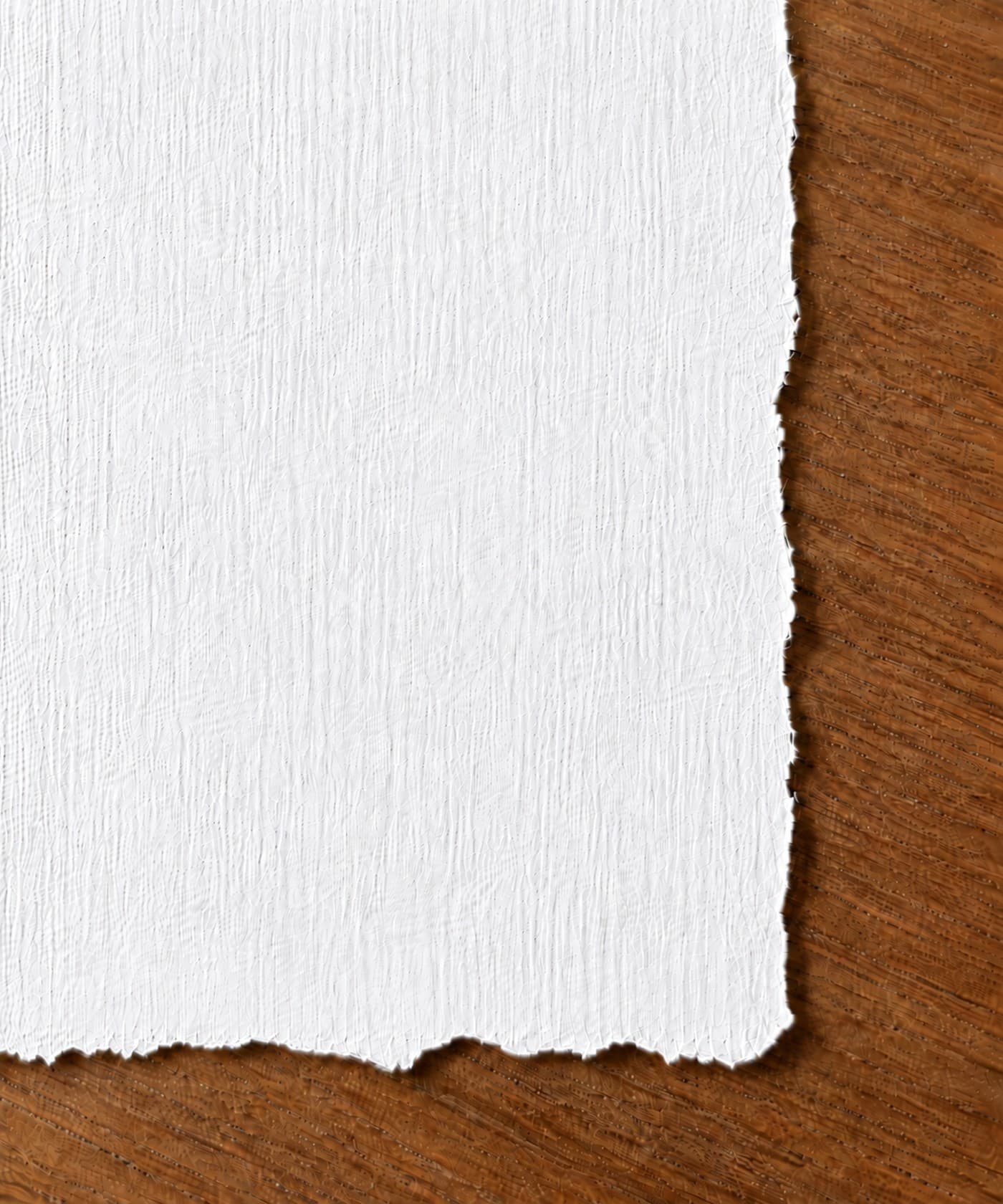 A close-up of a sheet of handmade 100% cotton fiber paper resting on a warm wood surface, its torn deckle edge feathering irregularly down the right side and across the bottom, with the fine fiber grain of the sheet visible in raking light.